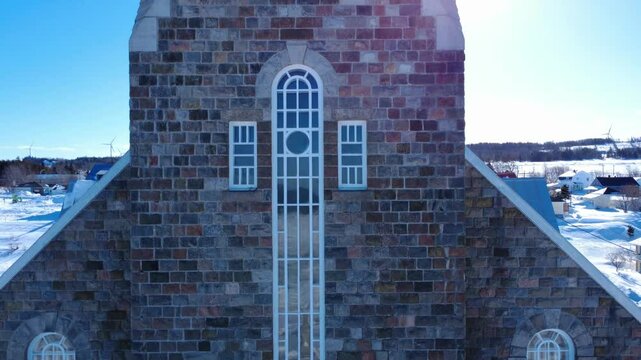 Drone video rotating from the stone brick facade to the metal cladding roof of the Assomption-de-Notre-Dame Church, in front of the St. Lawrence River frozen by the cold season. Baie-des-Sables QC CA