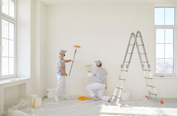 Two professional painters in uniform standing with their backs to camera and painting white wall with paint rollers. Male contractor workers engaged in ongoing renovation process in new apartment.