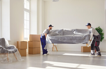 Two young moving service employees carrying heavy sofa together in the living room. Men workers from delivery service placing furniture in new house. Moving day and relocation concept.