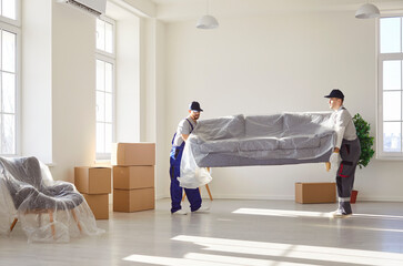 Two professional movers carrying wrapped sofa in new spacious apartment with cardboard boxes....