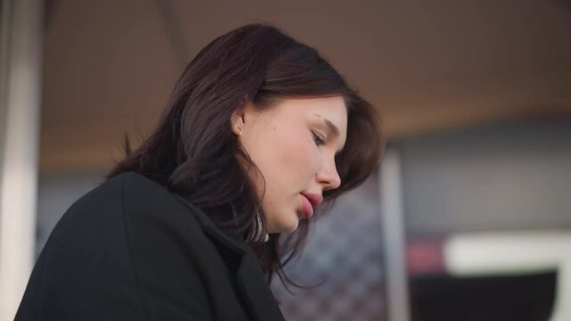 Close-up of white lady with pink lips muttering words softly, deeply focused on something in front of her, background features architectural structure with blurred details