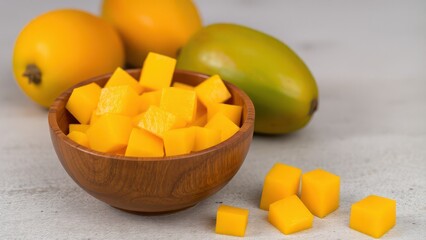 a wooden bowl filled with freshly cut mango cubes, with whole mangoes in the background and a few mango cubes placed on a textured surface.