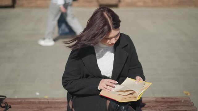 Woman in black coat sitting outdoors, engrossed in her book, she flips to a new page while her hair flutters in the wind, a passerby is seen in the background from behind