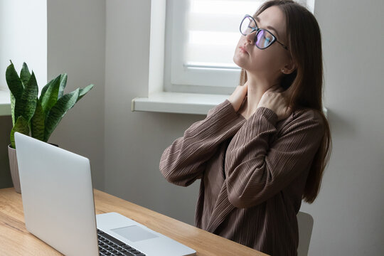 Overworked young woman, office worker, freelancer massaging neck and shoulder. Suffering from neck, shoulder pain while working on computer, laptop. Sedentary lifestyle.