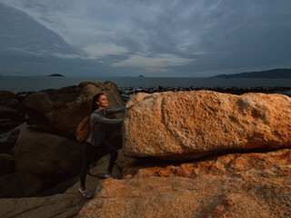 A young woman with a backpack climbing on rocky terrain by the sea, expressing determination and...