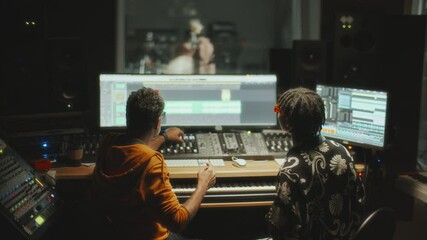 Two music producers working on audio tracks in professional audio studio with digital mixing equipment and large monitors, female singer recording vocals in booth behind glass