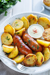 Roasted sausage with potatoes sprinkled with fresh herbs with addition aromatic sauce on white plate, close up view


