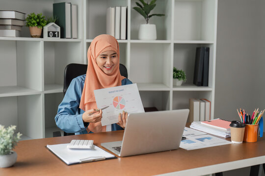 Muslim woman in hijab presenting a report with a pie chart during a video call in a modern office.
