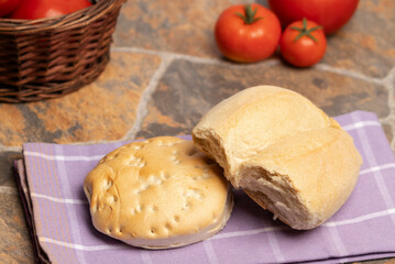Un par de panes de trigo, una marraqueta y una hallula sobre un paño de cocina, al fondo un canasto con tomates y tomates rojos y maduros