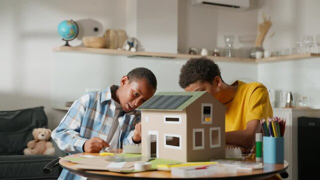 Younger brother turns on light inside solar-powered house model, illuminating the design, as both boys react with excitement and amazement. Scene emphasizes innovation and renewable energy education