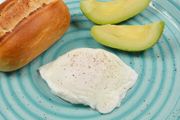 A top-down shot of a light blue plate with a bread roll, two slices of avocado, and a poached egg, presented as a simple breakfast meal