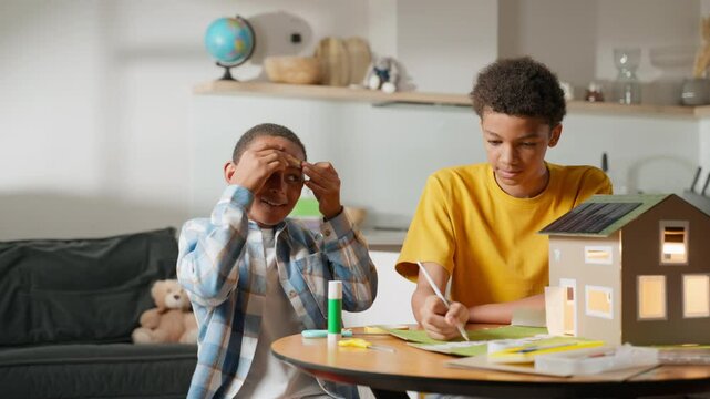 Younger brother playfully applies tape to his eyebrows, making older brother laugh. Lighthearted moment captures their close sibling bond, balancing fun with focus during school science project