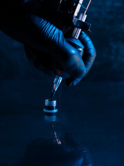 Close-up of a tattoo artist's gloved hand holding a tattoo machine, precisely dipping the needle into an ink cap. The scene is illuminated by moody blue lighting, emphasizing the artistry and precisio