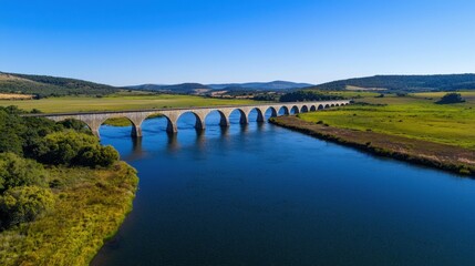 Fototapeta premium An expansive stone aqueduct gracefully crosses a calm river, framed by rolling hills and vibrant greenery. The bright blue sky completes this tranquil countryside view