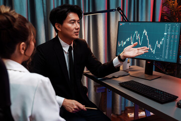 Asian businessman showing dynamic stock exchange currency rate statistic online to share with coworker, explaining to compare with high or low on computer screen at night neon modern office. Infobahn.