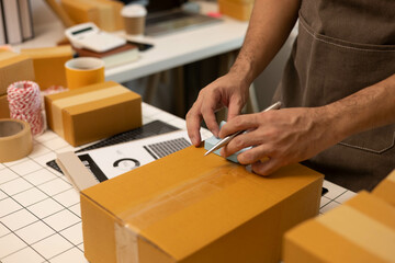 A man is sealing a brown cardboard box with tape. The box is on a table with a laptop and a stack...
