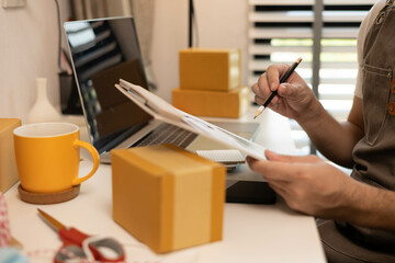 A man is sitting at a desk with a laptop and a clipboard. He is looking at a spreadsheet on the laptop and writing on the clipboard
