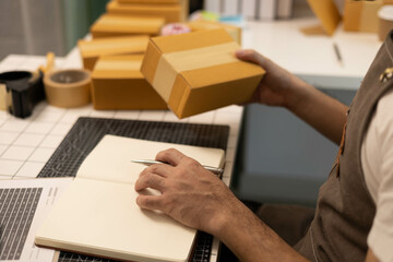 A man is writing in a notebook while holding a cardboard box. He is wearing an apron and he is working in a warehouse
