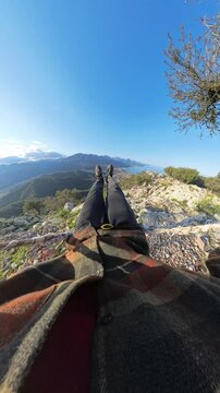 Vertical video. First-person view of a man swinging on a rope swing at the edge of a rocky cliff with a panoramic landscape of mountains, sea, and a coastal town in the background, captured with a wid