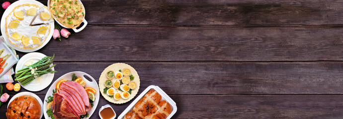 Classic Easter ham dinner. Overhead view corner border on a dark wood banner background with copy space. Ham, scalloped potatoes, vegetable side dishes, eggs, breads and lemon cake dessert.