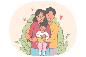 Happy family of mom and dad with little girl in arms, standing near bush and looking at camera