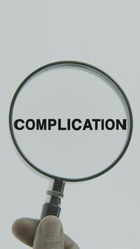 "Complication" text or word highlighted by a magnifying glass in a hand on a white background, symbolizing focus and analysis
