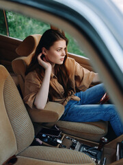 Young woman with long hair sitting in a vintage car, wearing a casual outfit, reflecting a serene mood with a green outdoor background.
