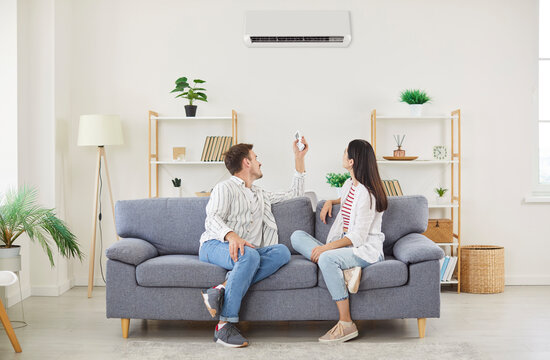Climate control, couple air conditioner fixing sitting relaxing on sofa holding controller, regulate climate at home using modern ac device, home ventilation to cool or warm new living room flat 