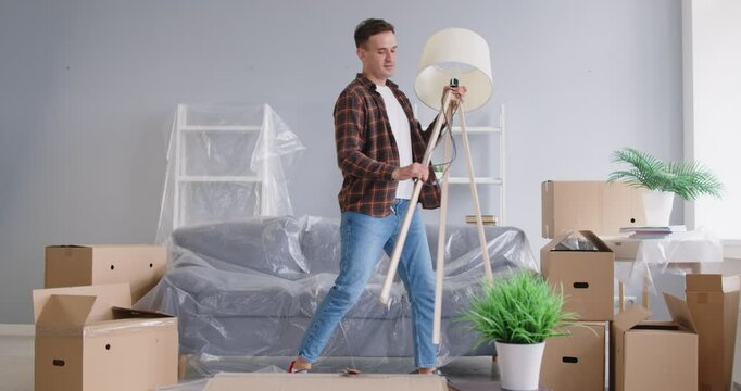 Man dances in room filled with cardboard boxes, showing his excitement during moving process. Man surrounded by cardboard boxes, furniture and flower pots is having fun on day of moving to new home.