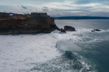 Atlantic ocean waves crashing over rocky coast during international surfers challenge at Farol da Nazaré in Nazare, Portugal on Moody cloudy February day.