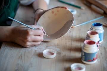 Unrecognizable ceramist applies flower paint to ceramic plate, making artistic author pattern. Creative occupation, handmade dishes, artisan clay production, unique craftsmanship, painted tableware