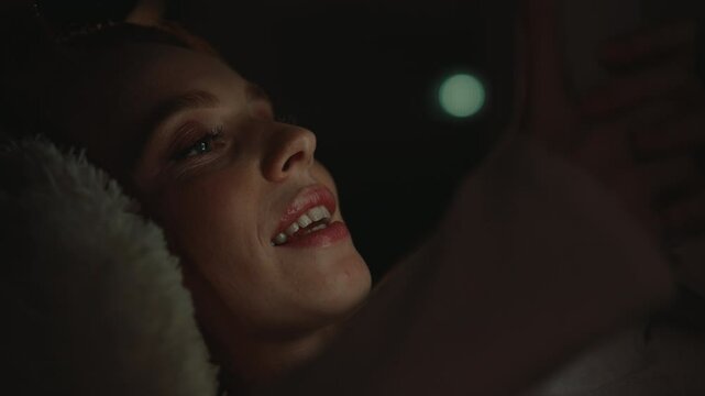Close-up face of happy young woman lying in bed at night, smiling using smartphone, with warm light glowing from screen, soft bokeh light on blurred background. Concept of social media addiction.