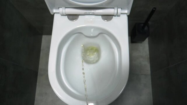 Bright yellow stream of urine pours into pristine white ceramic toilet, set in elegant modern bathroom with dark grey tiled walls and pristine interior design. First-person view.
