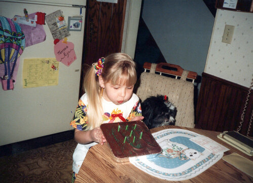 Circa 1990 - Child (girl) blows out candles on her chocolate birthday cake with her pet dog. Vintage archival photo