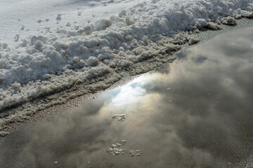 reflection of the sun on a puddle of water near a slush and snow bank of snow by the lake