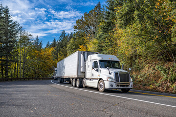 Low cab profile white big rig semi truck transporting cargo in refrigerator semi trailer running on the winding mountain road with autumn forest on the hillsides