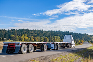 Powerful high cab white big rig semi truck tractor transporting two empty flat bed semi trailers running on the narrow winding road with autumn forest on the hill