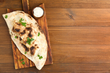 Board with tasty calzone, parsley and sauce on wooden table, top view. Space for text