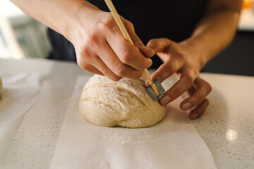 Woman hands skillfully score the surface of a round dough loaf, preparing sourdough bread for baking. The bright kitchen setting provides a warm atmosphere and fresh ingredients.