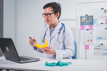 A male doctor stands holding a banana against a white background, promoting healthy eating.educates...