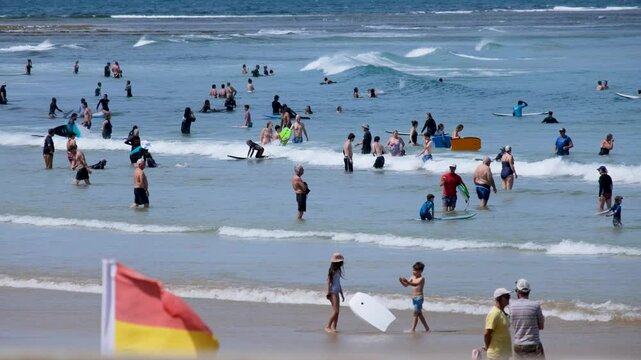 Crowded summer scene at Torquay surf beach shows swimmers, families, learner surfers, and beachgoers enjoying the gentle waves. It is a patrolled swimming area on Great Ocean Road in Australia