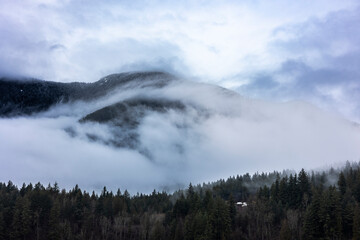 Foggy mountains in Hope, Canada, surrounded by trees on a cloudy gray day, contrasted with of blue sky. This picturesque scene captures the tranquility and beauty of nature in the Pacific North