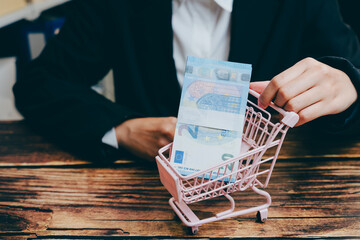 Businessman Holding Money in Mini Shopping Cart on Wooden Table, business concept