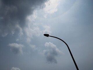 Silhouette of a curved streetlight against a cloudy sky with sunlight filtering through. A moody and dramatic scene showcasing urban minimalism and contrast between light and dark.
