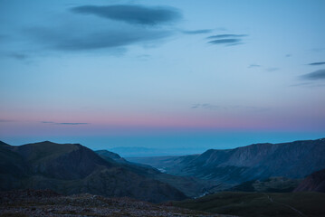 Top view from pass to wide alpine valley with long river against large mountain range silhouette with snow-capped peaks far away under lenticular clouds in sunset color sky. Big snowy ridge on horizon