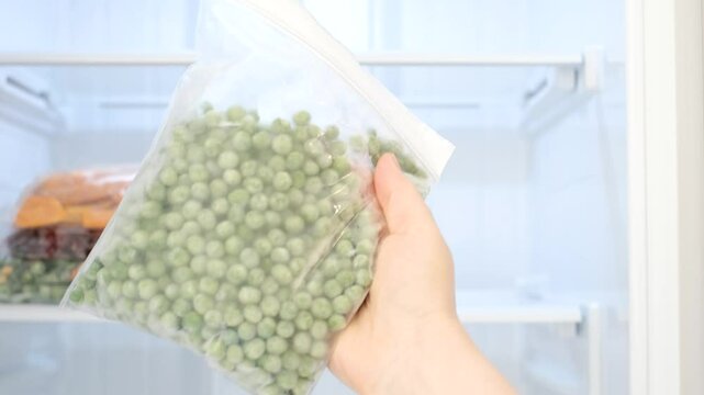 Female hand holding zip bag with frozen green peas against open freezer background.