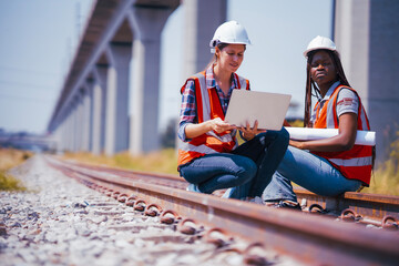 Black and White female Rail transportation engineers in safety vest and hardhat check the...