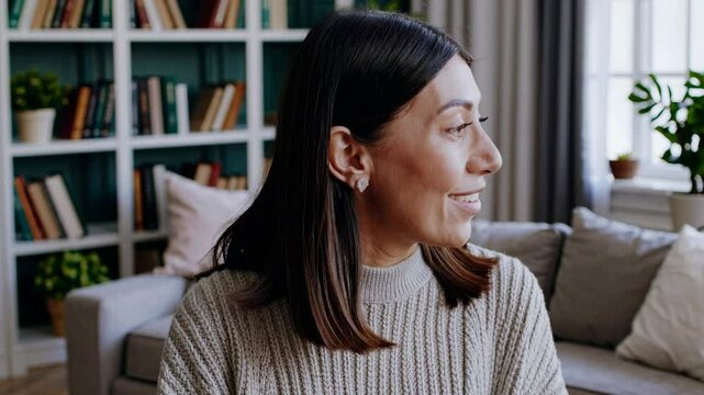 A woman smiles at the camera in a cozy living room setting, shot from a front-facing angle, ideal for a casual video call or vlog introduction.