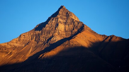 Naklejka premium A brown mountain stands tall in Glacier National Park as the sun rises, casting a warm glow on the peaks. A clear blue sky stretches above, creating a breathtaking landscape.