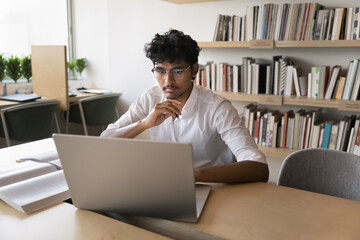 Concentrated Indian guy studying on laptop in college library, read article related to his diploma work, learning new educational platform, looks focused and interested. Modern tech, skills, knowledge